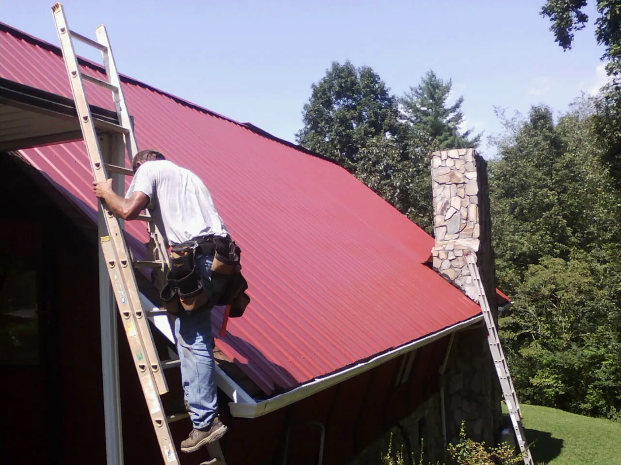 Roofer on a ladder beside a red metal roof and stone chimney — Soffit & Fascia Repair by Roofing Brothers in Thurmont